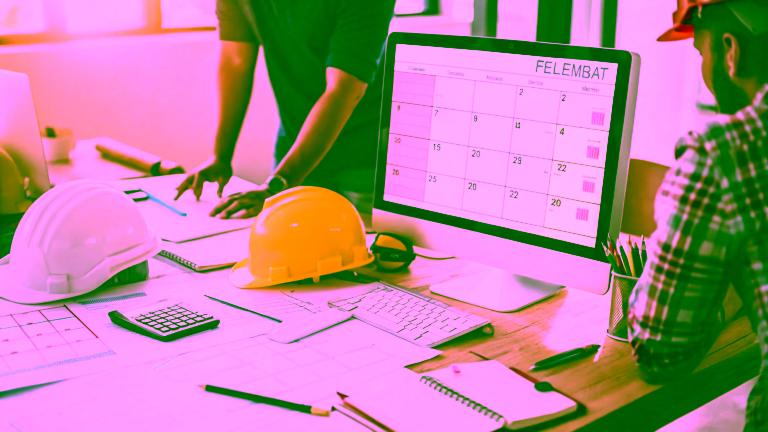 Construction site calendar on the computer, construction plans, helmets and office supplies on the desk.